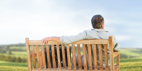 Ein älterer Mann sitzt auf einer Holzbank mit dem Rücken zur Kamera und betrachtet die grüne Hügellandschaft