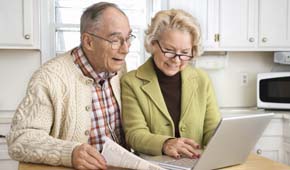 Ein älterer Mann und eine ältere Frau sitzen am Küchentisch und schauen begeistert auf den Monitor eines Laptops.