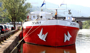 Das Ausstellungsschiff in rot und weiß auf einem Fluss. Es trägt normalerweise den Namen 'Jenny'.