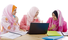 Drei muslimische Mädchen mit bunten Kleidern und Kopftüchern sitzen um einen Laptop und Schulunterlagen herum.