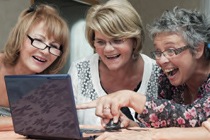 Das Foto zeigt drei ältere Damen, die sich lachend über einen Laptop beugen