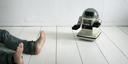 Das Bild zeigt einen kleinen Roboter, der neben den Füßen eines am Boden liegenden Menschen steht