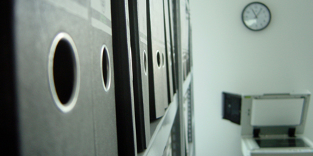 Das Bild zeigt einen Büroraum mit Aktenordnern im Regal und einem Kopiergerät