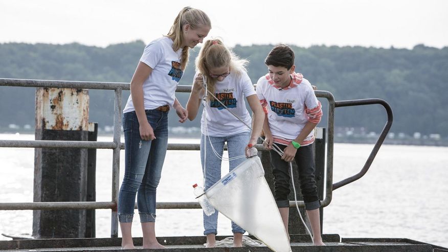 Foto, das zwei Schülerinnen und einen Schüler mit einem Mikroplastiksieb an einem Flussufer zeigt