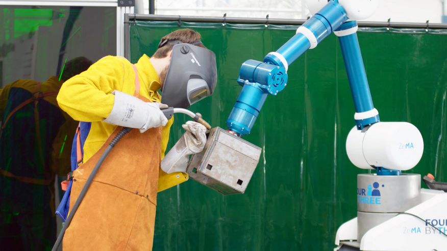 Zu sehen: Ein Roboterarm unterstütz einen Arbeiter beim Schweißen in dem er das Werkstück positioniert und hält