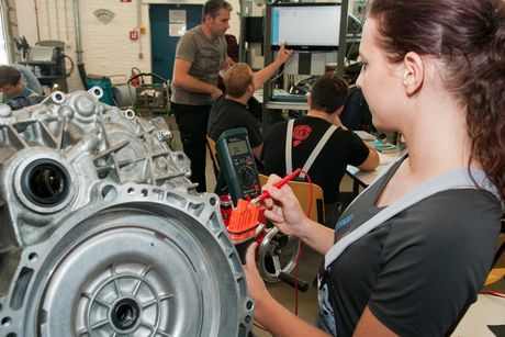 Im Vordergrund übt eine Kfz-Mechatronikerin in Ausbildung an einem Motor. Im Hintergrund lesen Auszubildene Daten von einem Bildschirm ab.