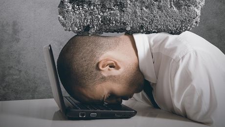 Symbolbild, ein Mensch liegt mit seiner Stirn auf einem Laptop. Auf seinem Kopf liegt ein schwerer Stein. Dies soll den digitalen Stress symbolisieren.