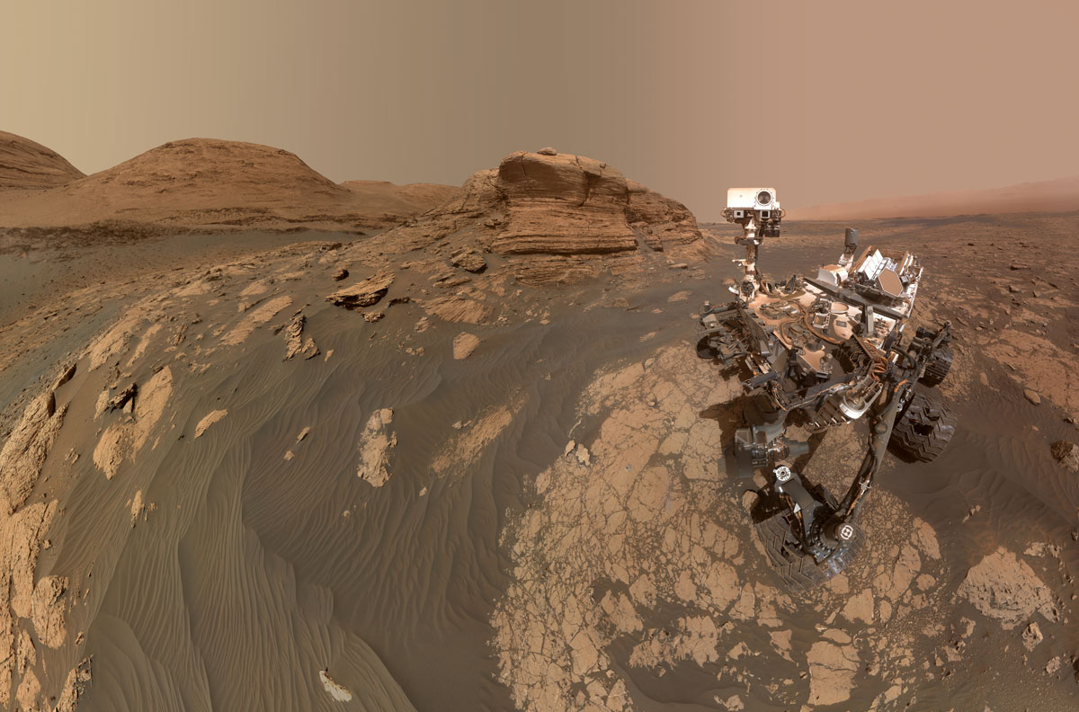Marsrover Curiosity auf dem Mars. Marsboden und -himmel sind rötlich gefärbt. Im Hintergrund sind Berge zu sehen. 