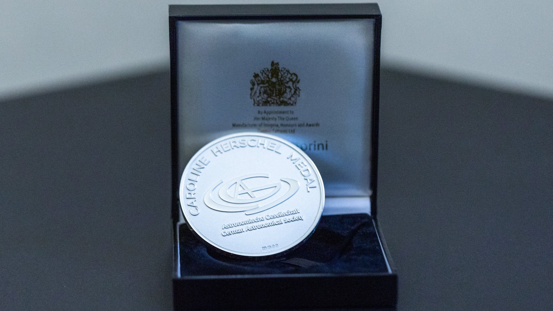 Eine silberne Medaille mit der Aufschrift „Caroline Herschel Medal, Astronomische Gesellschaft, German Astronomical Society“, die in einem geöffneten Kästchen liegt.