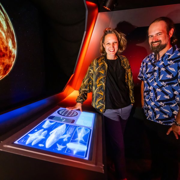 Eine Frau und ein Mann im Cockpit neben einem großen Display, der einen Planeten zeigt.