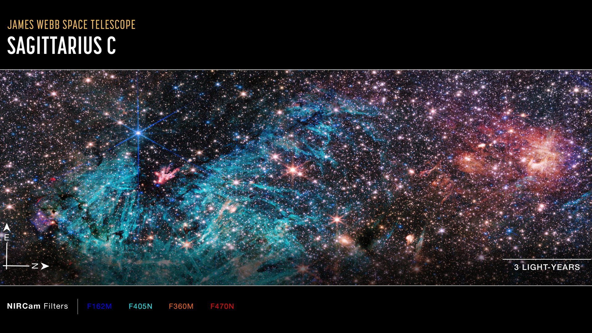 Auf schwarzem Hintergrund leuchten viele Sterne in verschiedenen Farbtönen. In Form eines dicken U zieht sich eine hellblau leuchtende Wolke durch die untere Hälfte des Bildes, in der dunkle Flecken und kurze, helle Linien auszumachen sind. In der dunklen Delle über dem U leuchtet ein kleiner, rosa Fleck mit weißen Punkten. 