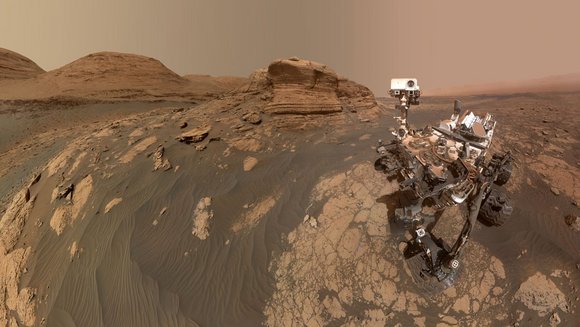 Marsrover Curiosity auf dem Mars. Marsboden und -himmel sind rötlich gefärbt. Im Hintergrund sind Berge zu sehen. 