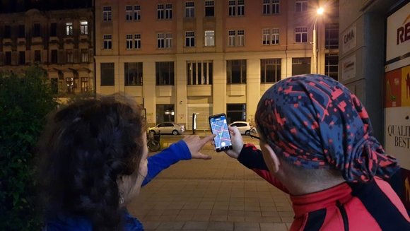 Ein Mann und eine Frau stehen bei Nacht vor einem Haus mit einer Straßenlaterne. Sie stehen mit dem Rücken zur Kamera und halten ein Handy mit der Nachtlicht-App in der Hand. 