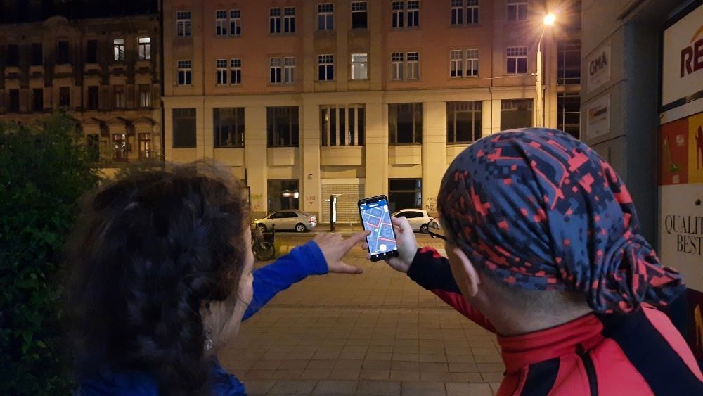 Ein Mann und eine Frau stehen bei Nacht vor einem Haus mit einer Straßenlaterne. Sie stehen mit dem Rücken zur Kamera und halten ein Handy mit der Nachtlicht-App in der Hand. 