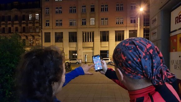 Ein Mann und eine Frau stehen bei Nacht vor einem Haus mit einer Straßenlaterne. Sie stehen mit dem Rücken zur Kamera und halten ein Handy mit der Nachtlicht-App in der Hand. 