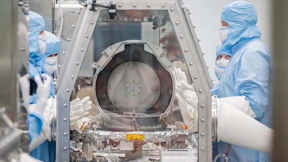 Menschen in blauen Schutzanzügen stehen um einen Kanister, der sich in einem Glasbehälter befindet.