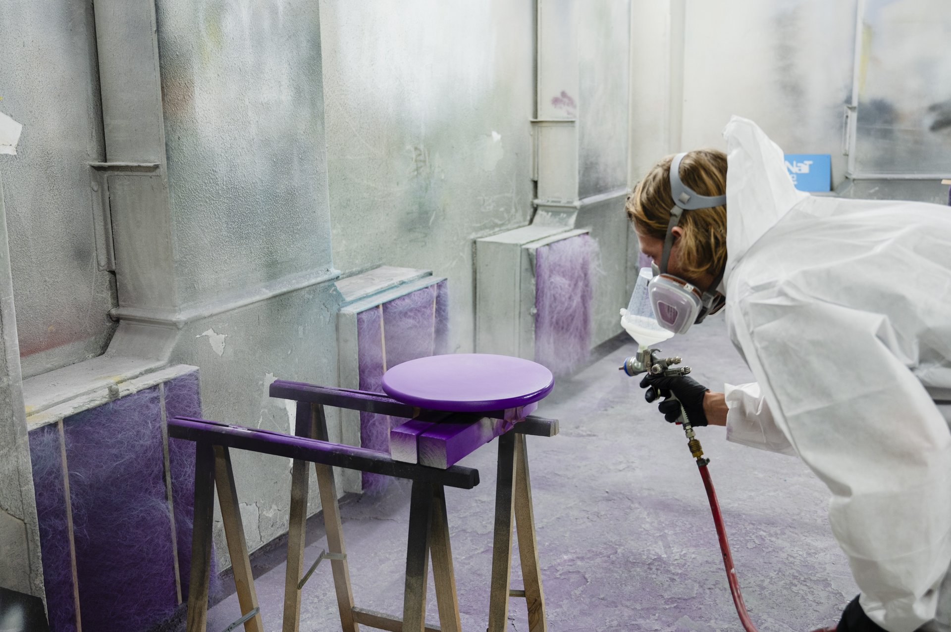 Eine Person sprüht lila Farbe auf eine runde Platte. Sie trägt einen Maleranzug und eine Schutzmaske. 