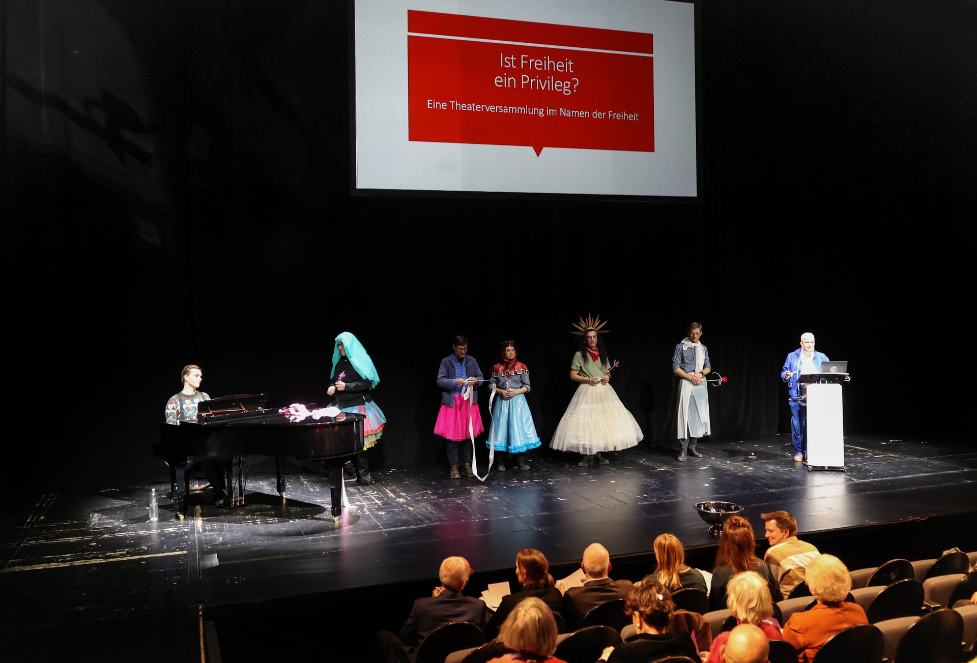 Die Darstellenden und Sibylle Peters auf der Bühne, darüber der Veranstaltungstitel auf einer Leinwand.