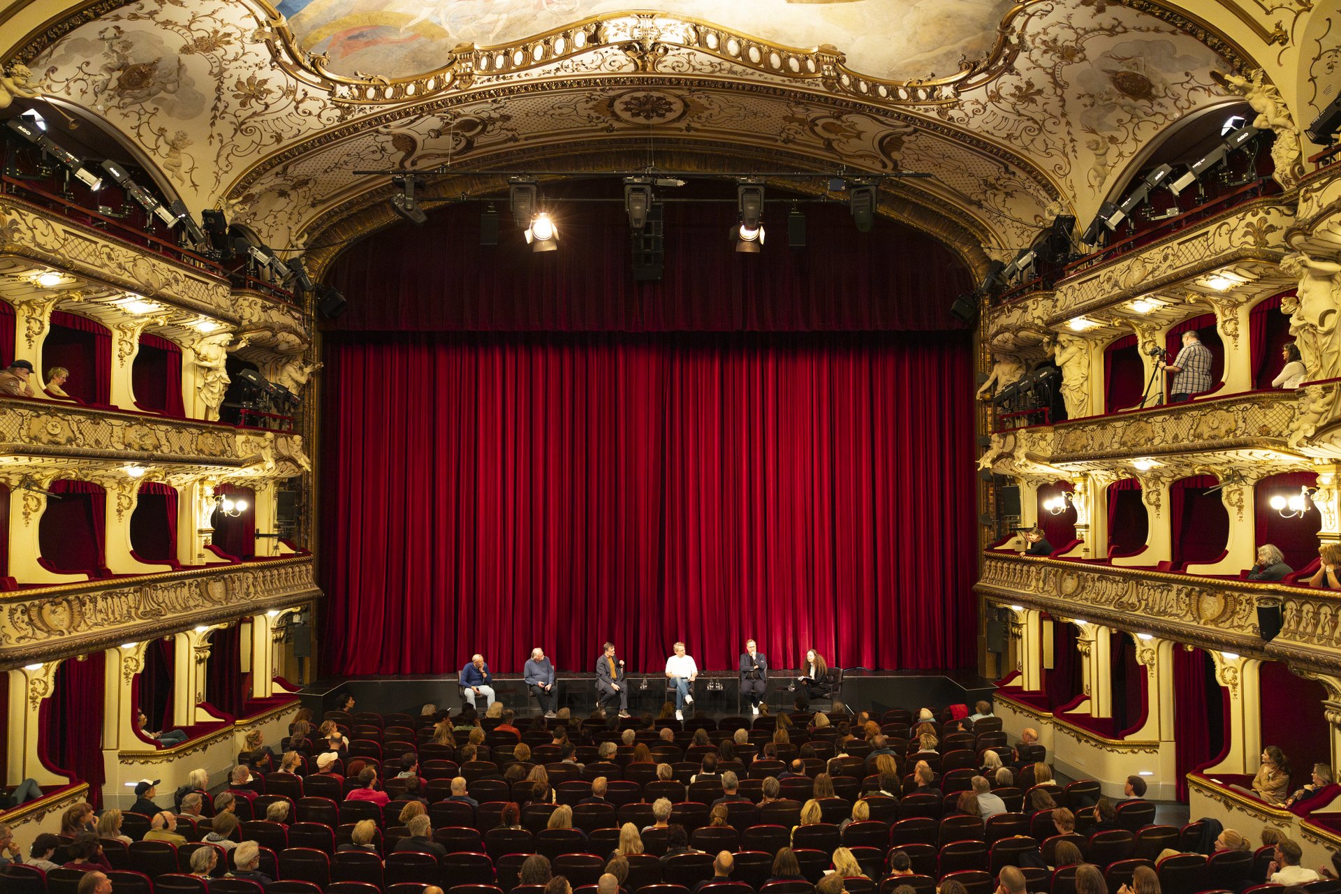 Totale der Theaterbühne des SchauSpielHauses. Vor dem roten Vorhang sitzt das Panel, bestehend aus fünf Personen. 