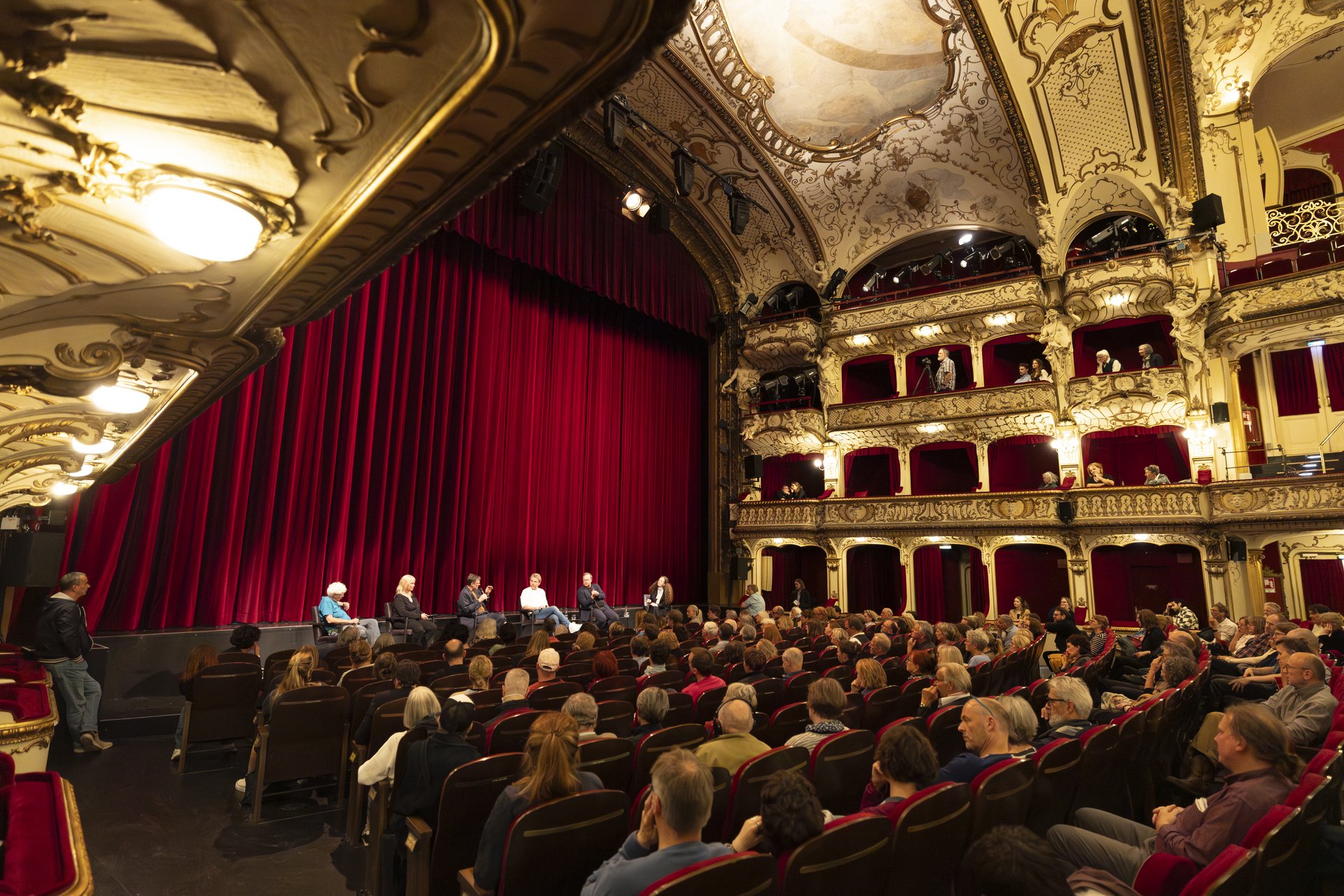 Die Theaterbühne des SchauSpielHauses von der linken Seite. Vor der Bühne und an der rechten Seite sind Reihen mit vielen Zuschauenden gefüllt. 