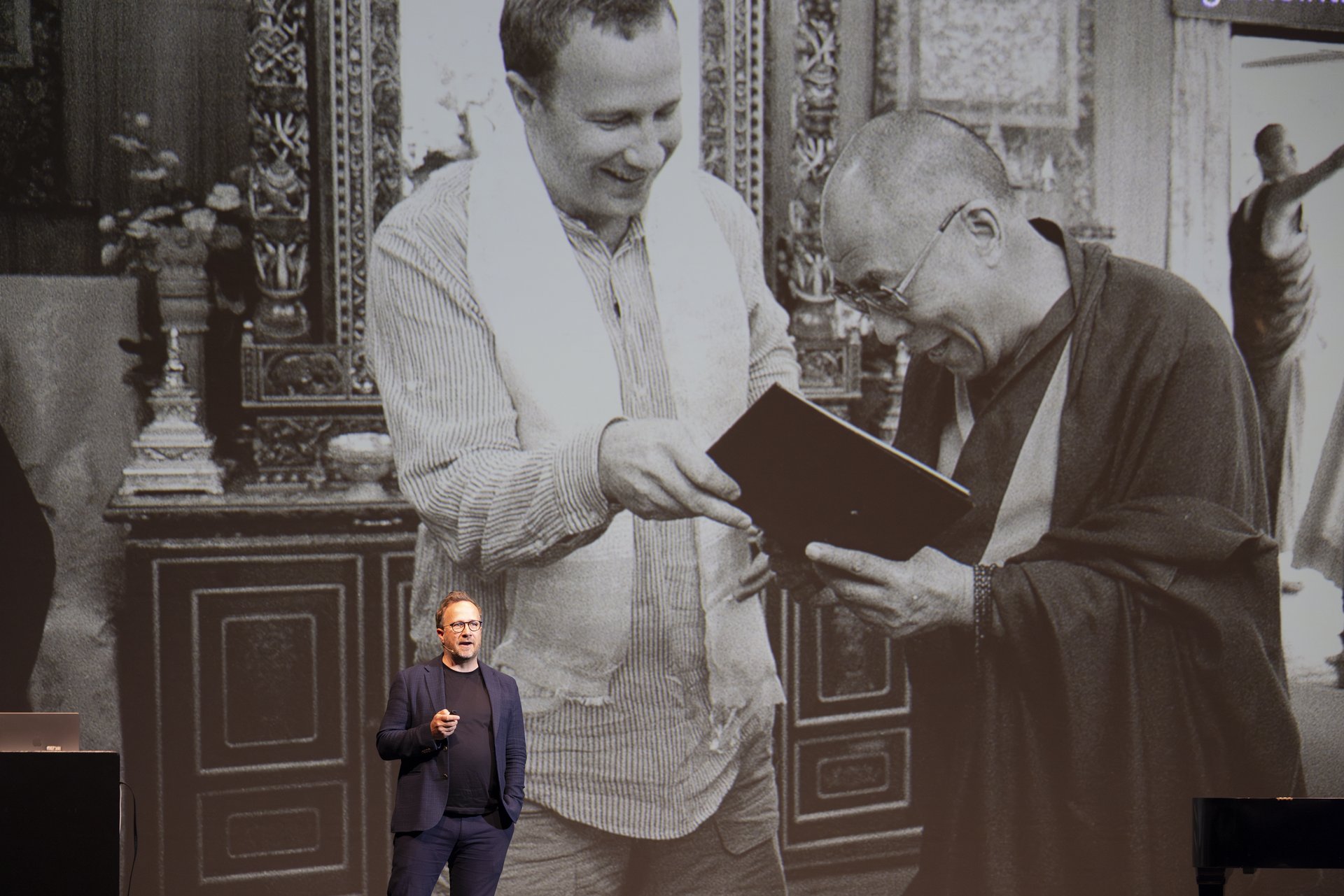 Andri Snær Magnason inszeniert auf der Bühne eine Lecture Performance. Im Hintergrund wird ein schwarz weiß Bild von zwei Männern auf die Wand projiziert, die ein Buch in der Hand halten und sich darüber unterhalten. 