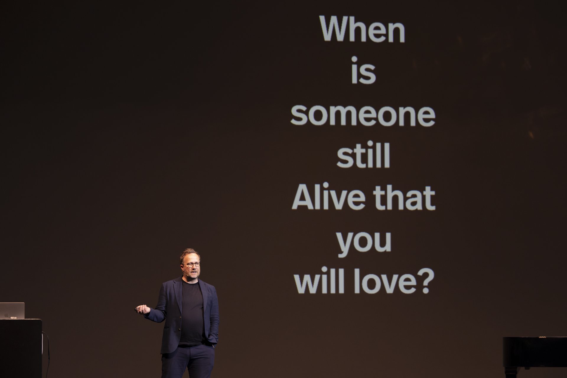 Andri Snær Magnason auf der Bühne. Im Hintergrund wird die Leitfrage seiner Lecture Performance auf die Wand projiziert: „When is someone still alive that you love”.
