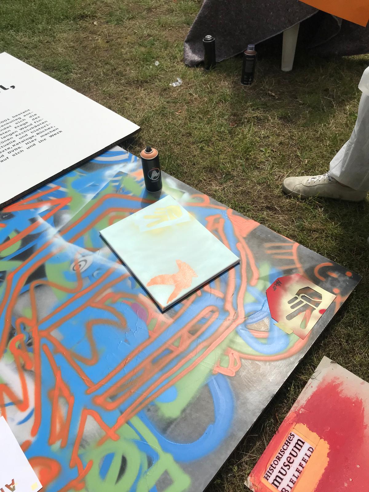 Bunte Leinwand mit abstrakten Graffiti-Mustern. Auf der Leinwand ist liegen eine Sprühdose und eine Vogel-Schablone.