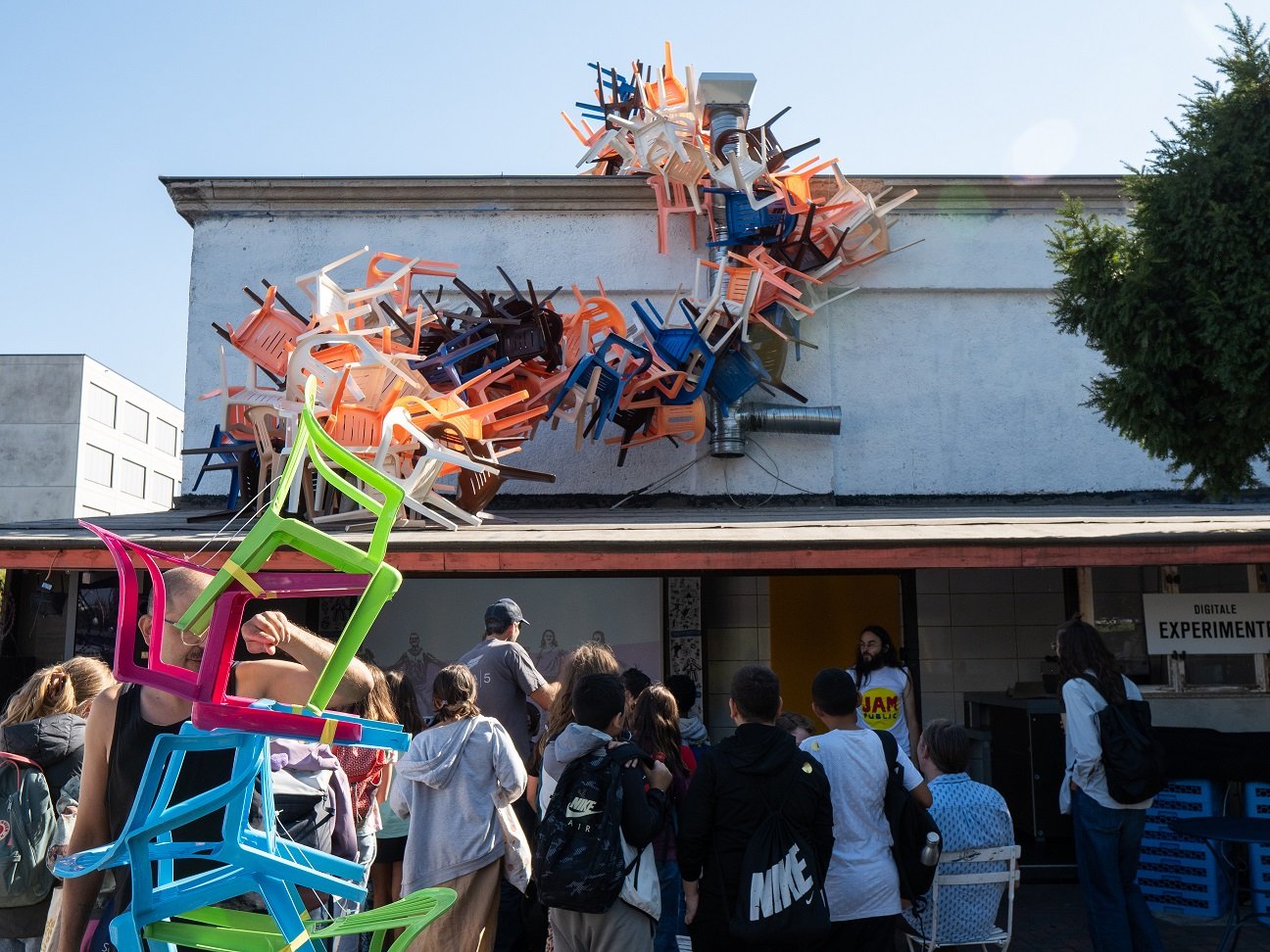 Ein Stapel Plastikstühle ragt über einer Gruppe Menschen und einem Hausdach.