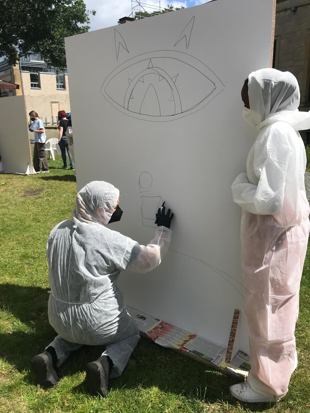 Zwei Teilnehmende in weißer Schutzkleidung skizzieren ein Design auf einer großen Leinwand.