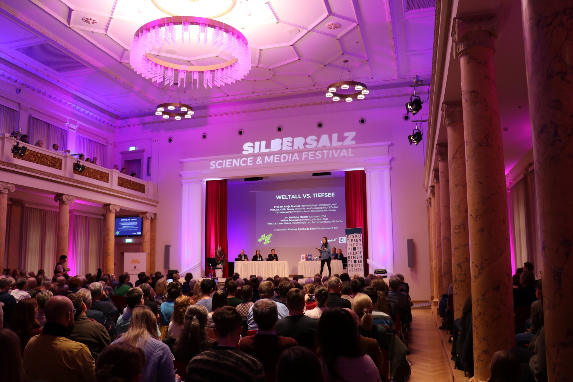 Projektbild Wissenschaft kontrovers: Foto einer Frau auf der Bühne eines gefüllten Saals. Auf der Bühne sitzen sechs weitere Menschen. Die Veranstaltung ist überschrieben mit "Silbersalz. Science and Media Festival".