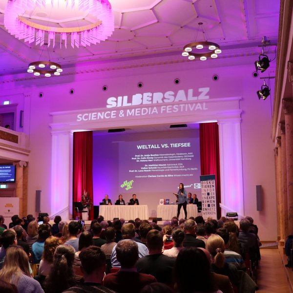 Projektbild Wissenschaft kontrovers: Foto einer Frau auf der Bühne eines gefüllten Saals. Auf der Bühne sitzen sechs weitere Menschen. Die Veranstaltung ist überschrieben mit "Silbersalz. Science and Media Festival".