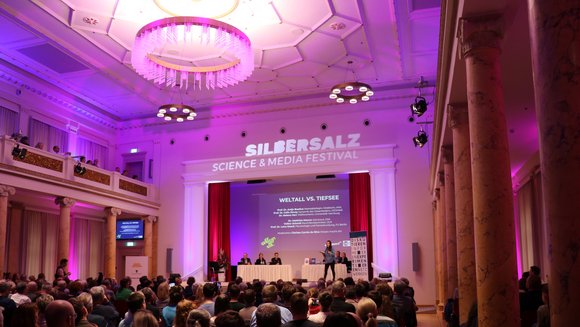 Projektbild Wissenschaft kontrovers: Foto einer Frau auf der Bühne eines gefüllten Saals. Auf der Bühne sitzen sechs weitere Menschen. Die Veranstaltung ist überschrieben mit "Silbersalz. Science and Media Festival".