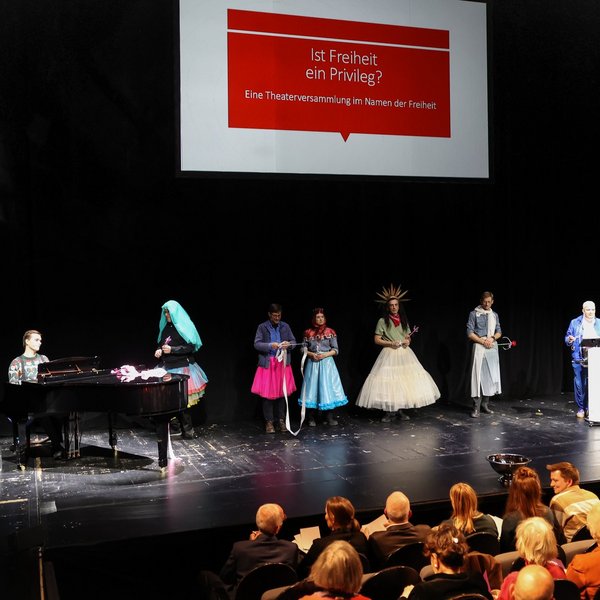 Die Darstellenden und Sibylle Peters auf der Bühne, darüber der Veranstaltungstitel auf einer Leinwand.