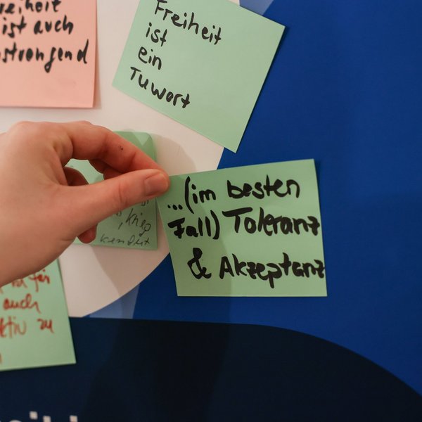 Eine Person klebt einen Zettel mit der Aufschrift „Im besten Fall Toleranz & Akzeptanz“ auf eine Tafel.