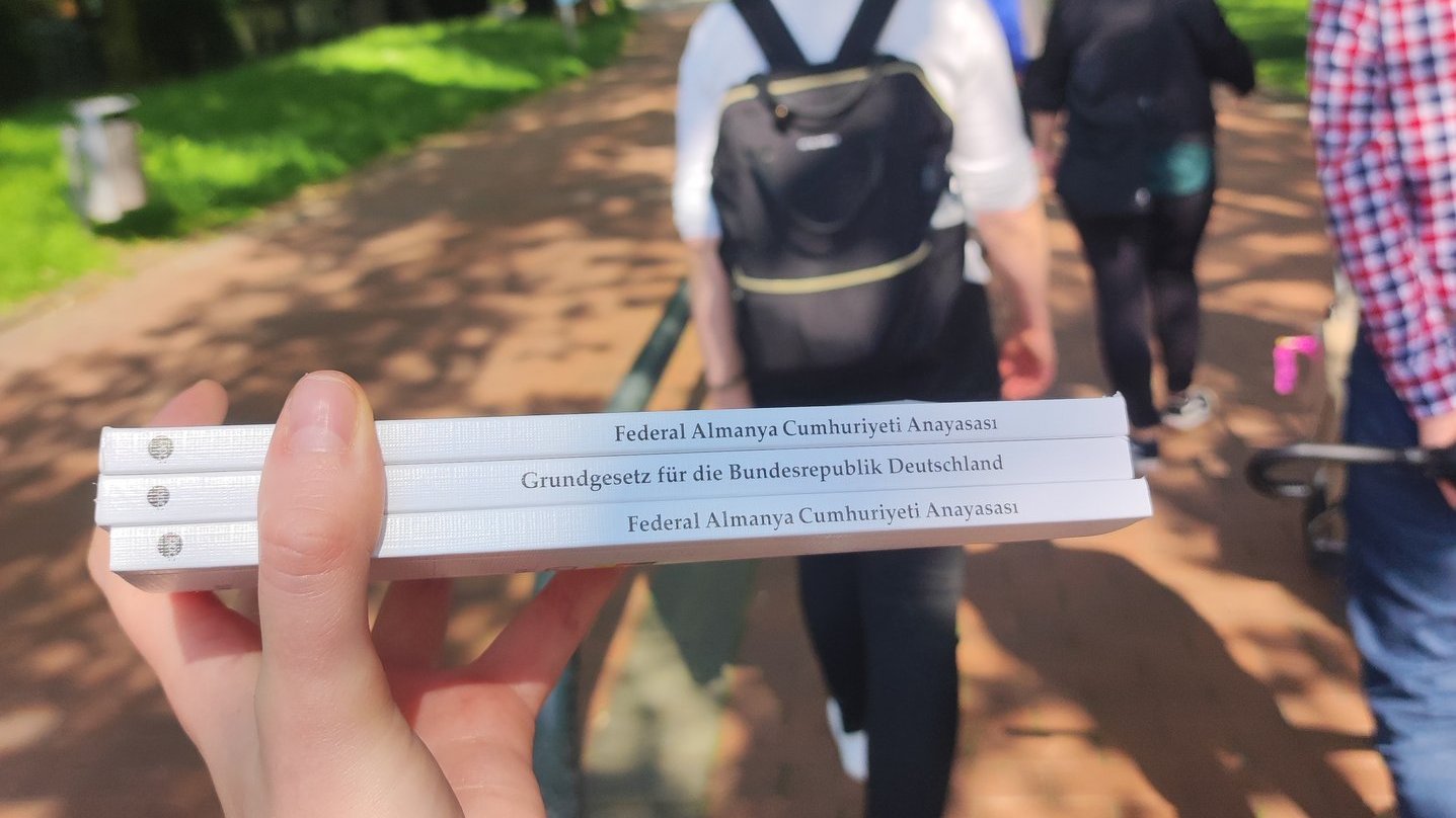 Foto vom Grundgesetz in mehreren Ausgaben, im Hintergrund spazieren Menschen.