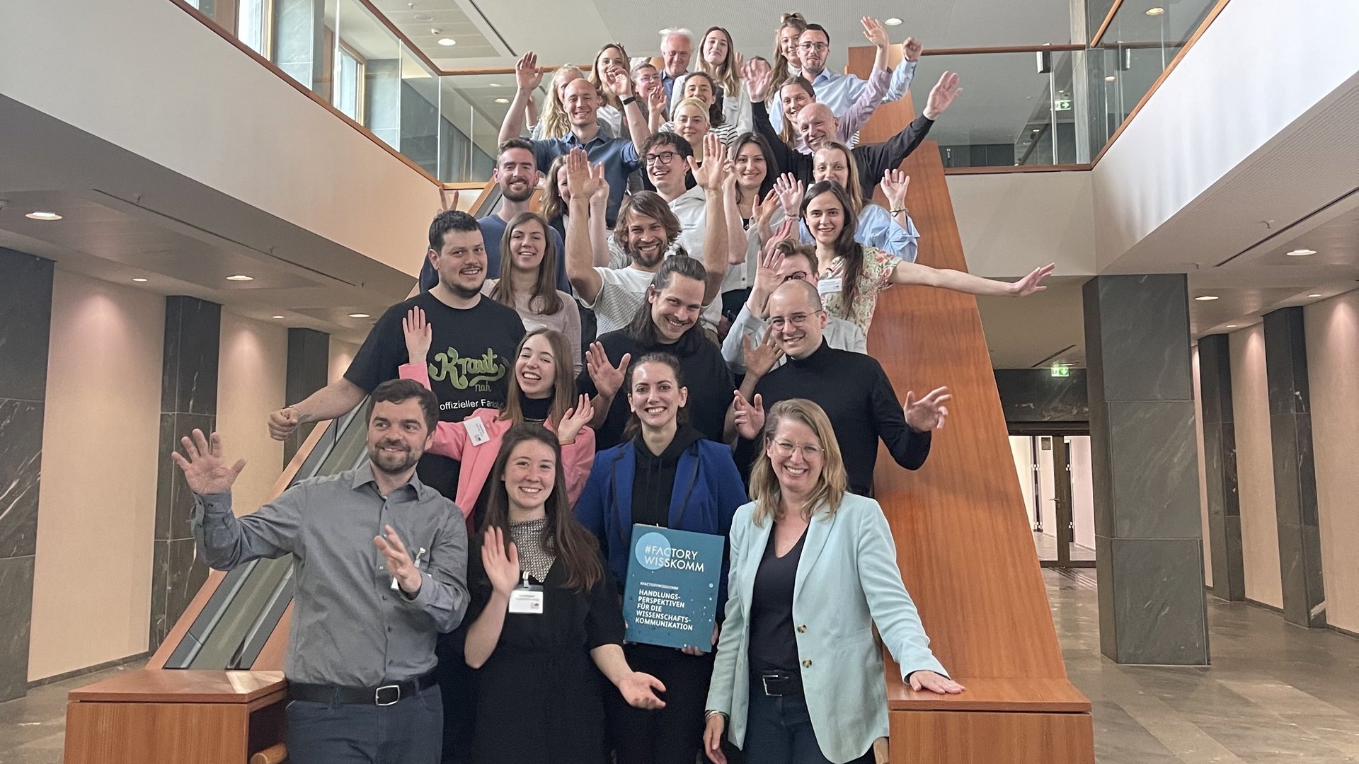 Projektbild Hochschulwettbewerb: Foto mit etwa 25 Menschen, die von einer Treppe winken. Eine Frau in der ersten Reihe hält eine Mappe mit der Aufschrift "#FACTORY WISSKOMM".