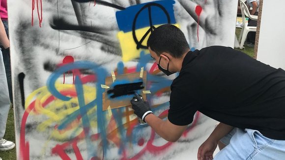 Eine teilnehmende Person gestaltet eine große Leinwand mit Graffiti. Auf ihr steht groß "PEACE" in gelb und blau. Unten ist die ukrainische Flagge zu sehen.