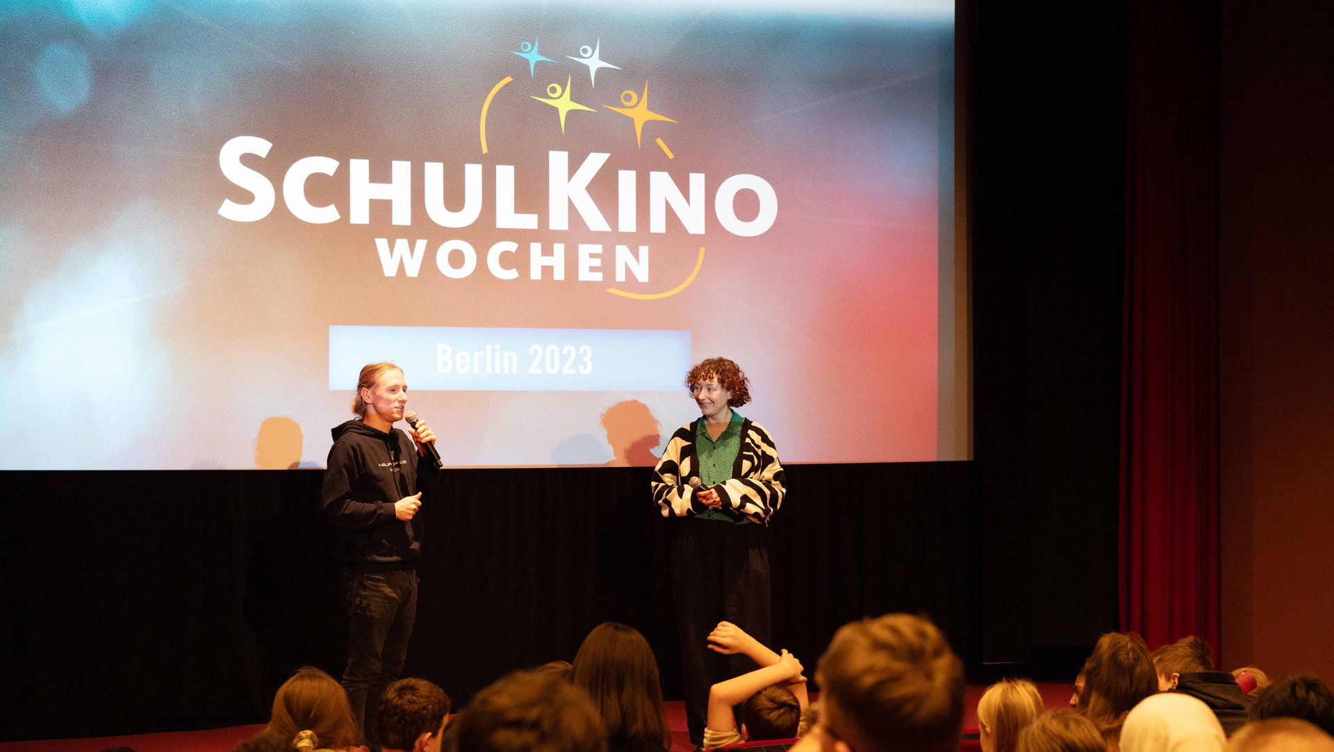 Projektbild SchulKinoWochen: Foto von zwei jungen Erwachsenen, die beide ein Mikrofon halten. Sie stehen vor einer Gruppe Schülerinnen und Schüler in einem Kinosaal und unterhalten sich.