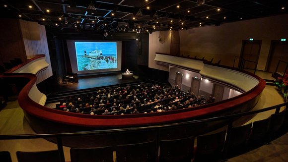 Der Saal im Volkstheater mit Publikum, auf die Leinwand ist ein Schiff am Hafen projiziert.