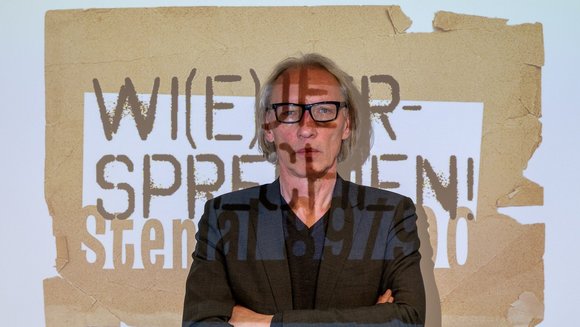 Prof. Dr. habil. Günter Mey steht vor einer weißen Wand, auf die der Schriftzug "Wi(e)versprechen! Stendal 89/90" projiziert wird.