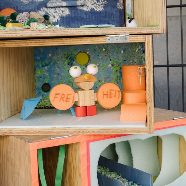 Mehrere Holzkisten, in der Mitte steht eine Figur mit der Beschriftung „Freiheit“.