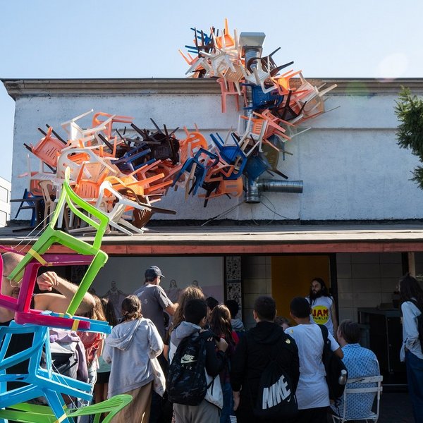 Ein Stapel Plastikstühle ragt über einer Gruppe Menschen und einem Hausdach.