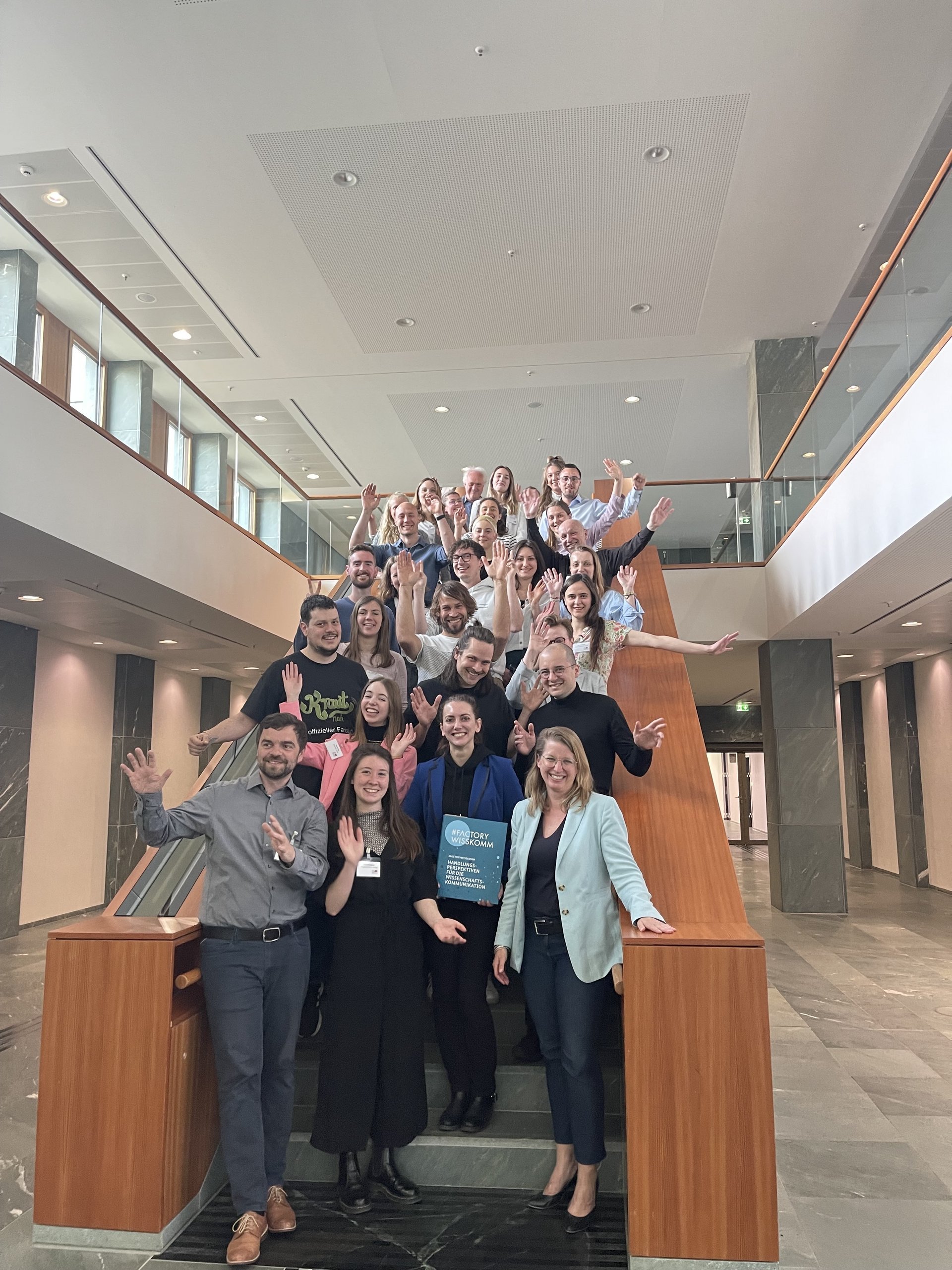 Projektbild Hochschulwettbewerb: Foto mit etwa 25 Menschen, die von einer Treppe winken. Eine Frau in der ersten Reihe hält eine Mappe mit der Aufschrift "#FACTORY WISSKOMM".