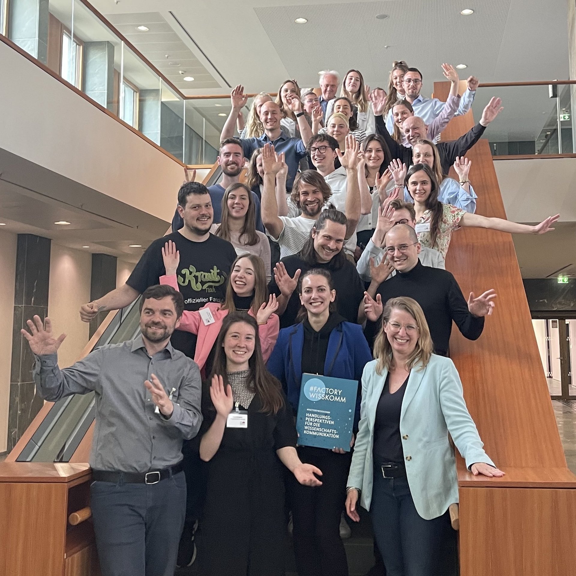 Projektbild Hochschulwettbewerb: Foto mit etwa 25 Menschen, die von einer Treppe winken. Eine Frau in der ersten Reihe hält eine Mappe mit der Aufschrift "#FACTORY WISSKOMM".