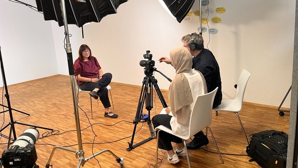 Eine Frau wird von zwei Gesprächspartner:innen interviewt und dabei gefilmt.