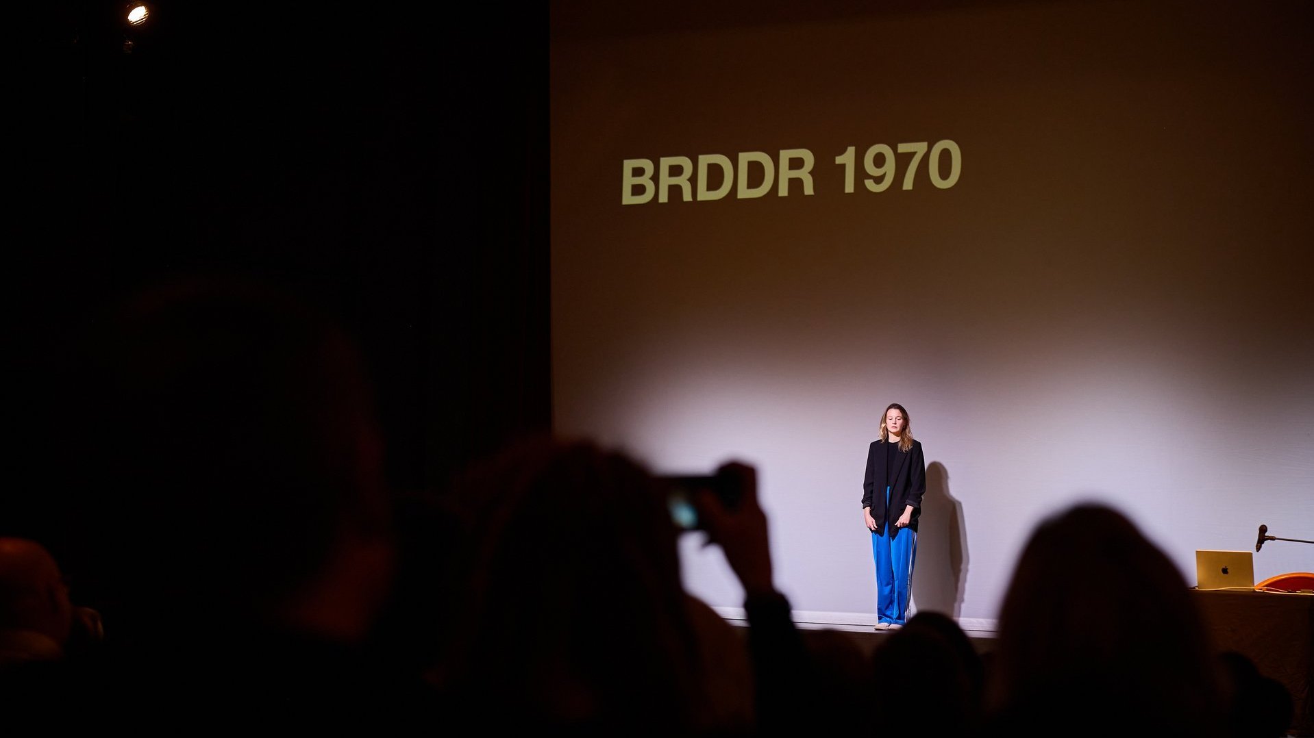 Eine junge Frau vor Publikum, hinter ihr steht „BRDDR 1970“.