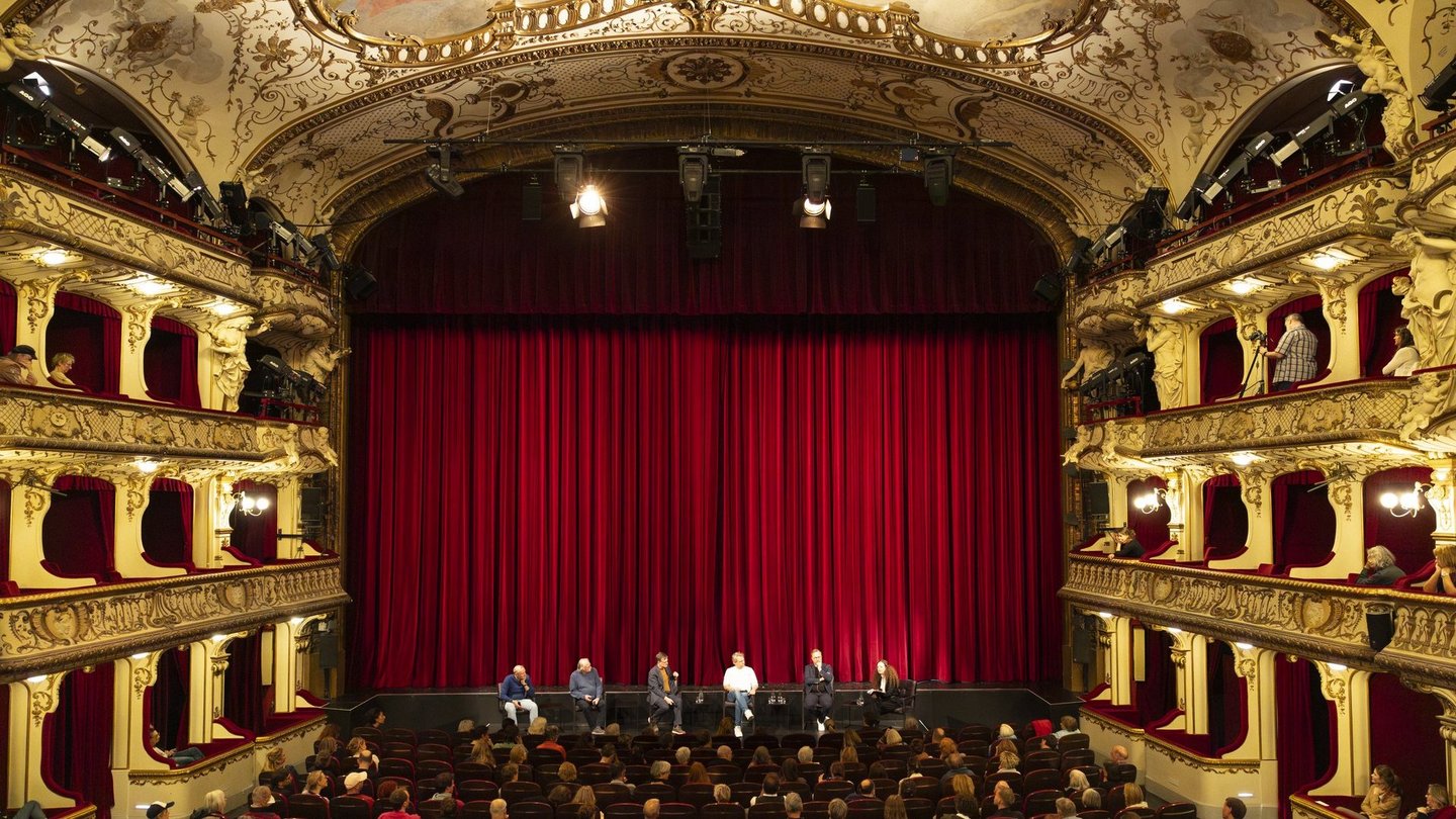 Totale der Theaterbühne des SchauSpielHauses. Vor dem roten Vorhang sitzt das Panel, bestehend aus fünf Personen. 