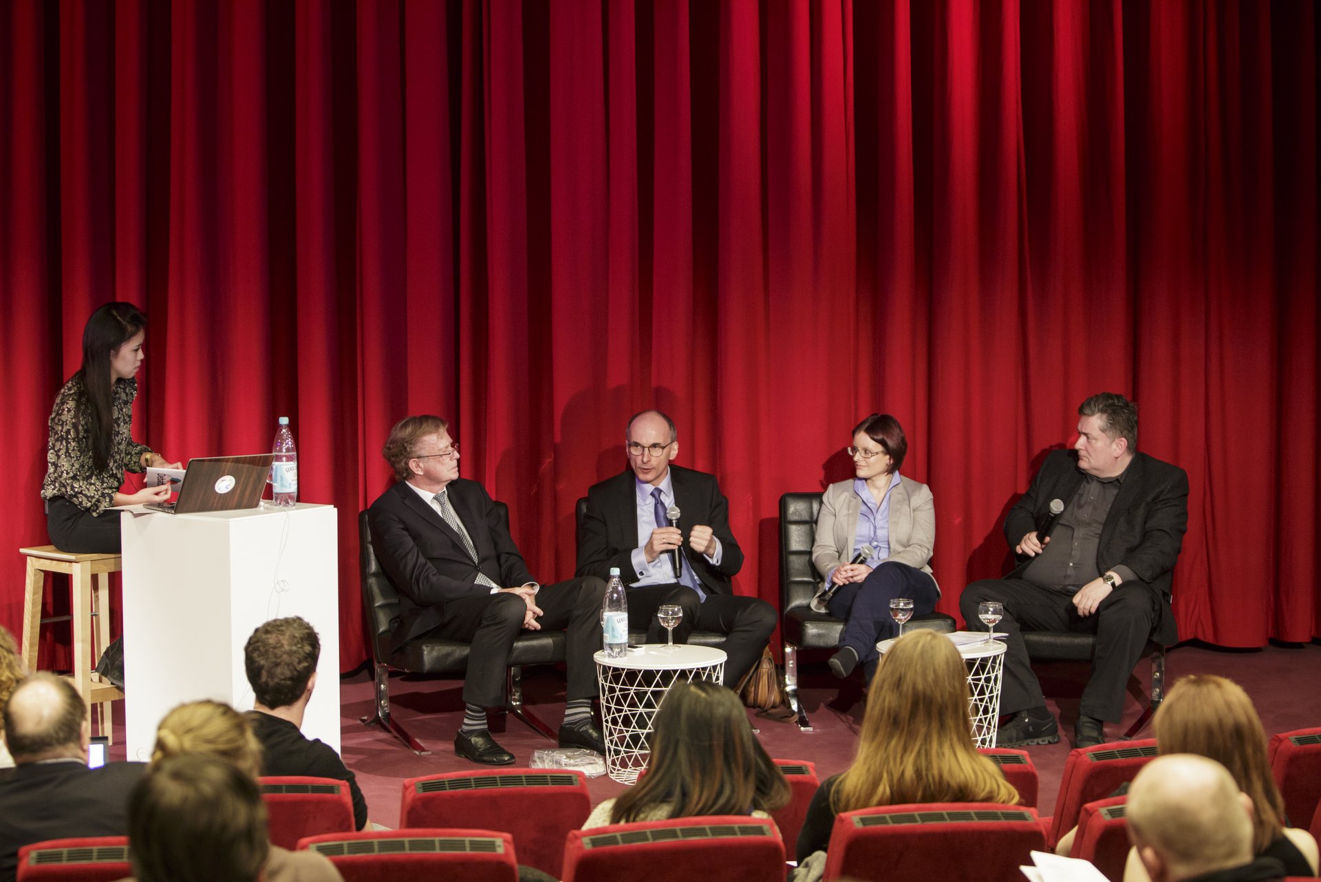Vier Personen debattieren auf einer Bühne mit rotem Vorhang im Hintergrund