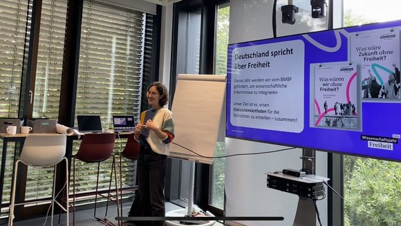 Eine junge Frau moderiert einen Workshop neben einem Bildschirm.