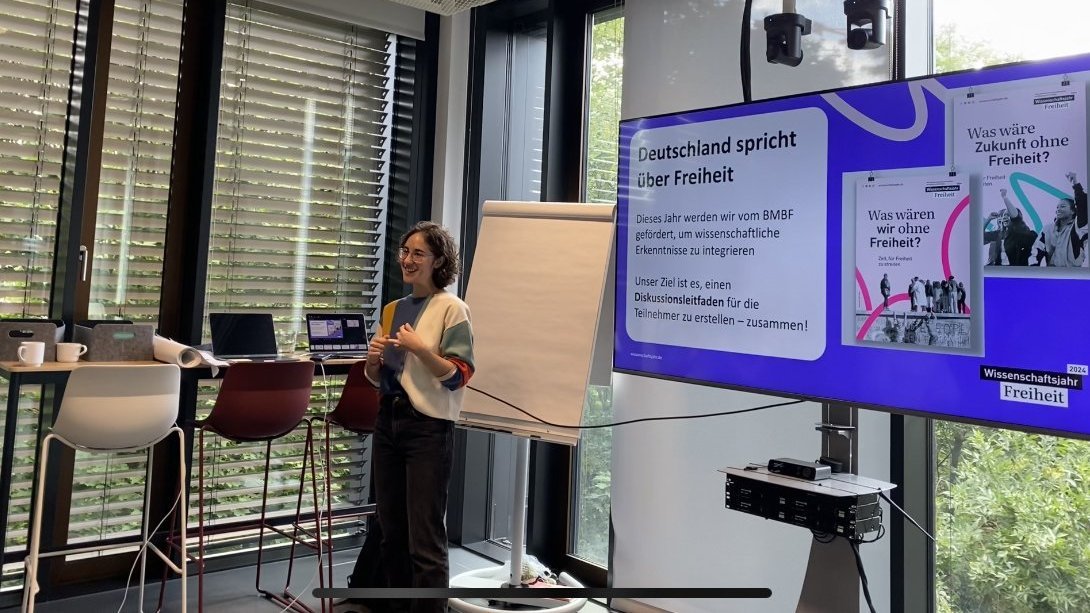 Eine junge Frau moderiert einen Workshop neben einem Bildschirm.